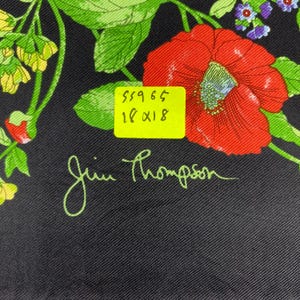Puede incluir: Pa&ntilde;uelo de seda negro con un dise&ntilde;o floral que presenta una gran amapola roja, hojas verdes y flores moradas m&aacute;s peque&ntilde;as. El pa&ntilde;uelo tiene la firma "Jim Thompson" en escritura verde y una etiqueta amarilla con n&uacute;meros.