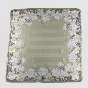 Puede incluir: Un pañuelo cuadrado con un diseño floral. El pañuelo presenta un fondo verde claro con rayas horizontales y un borde de lirios blancos. Los bordes son de un tono verde más oscuro.