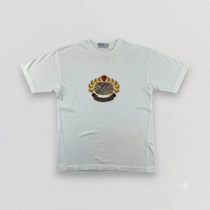 May include: A white short-sleeve t-shirt featuring a central embroidered crest logo. The design includes a golden laurel wreath surrounding a knight on horseback, with a dark blue banner below displaying the text BURBERRYS in white letters.