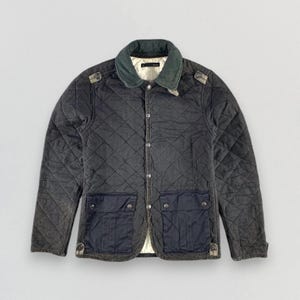 Puede incluir: Una chaqueta acolchada de color verde oscuro con cuello de pana y dos bolsillos grandes con solapa. La chaqueta tiene cierre de botones y un aspecto desgastado.