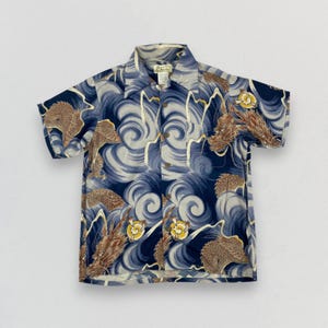 Puede incluir: Camisa de manga corta con un patrón de olas azules y blancas e ilustraciones de dragones marrones. La camisa tiene un cuello clásico y un corte relajado.