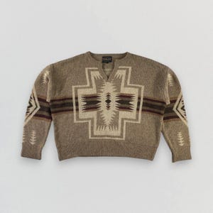 Op de afbeelding: Een bruine v-hals trui met een geometrisch patroon in beige, bruin en bordeaux. De trui heeft een centraal kruisvormig ontwerp en patronen op de mouwen. De merknaam is zichtbaar op de halslijn.