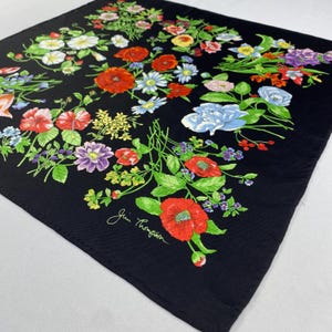 Puede incluir: Pa&ntilde;uelo cuadrado negro con un vibrante dise&ntilde;o floral. El pa&ntilde;uelo presenta una variedad de flores coloridas, incluyendo amapolas rojas, acianos azules y margaritas blancas, sobre un fondo negro. El pa&ntilde;uelo tiene la firma "Jim Thompson" en la esquina.