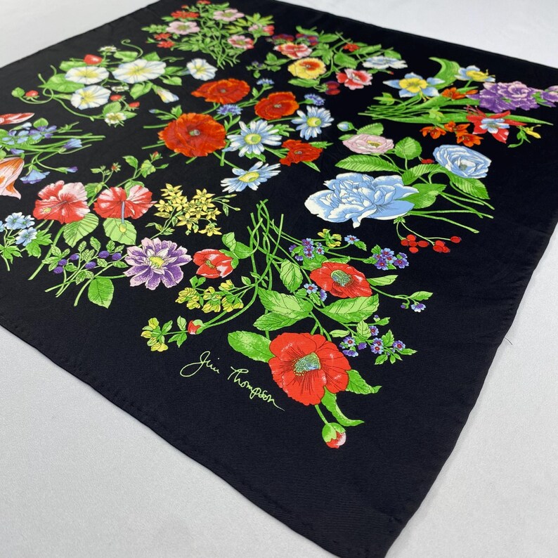Puede incluir: Pa&ntilde;uelo cuadrado negro con un vibrante dise&ntilde;o floral. El pa&ntilde;uelo presenta una variedad de flores coloridas, incluyendo amapolas rojas, acianos azules y margaritas blancas, sobre un fondo negro. El pa&ntilde;uelo tiene la firma "Jim Thompson" en la esquina.