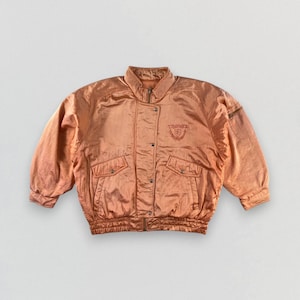 Puede incluir: Una chaqueta bomber vintage de satén naranja con cierre de cremallera y un logotipo bordado en el pecho. La chaqueta tiene cintura y puños de canalé.