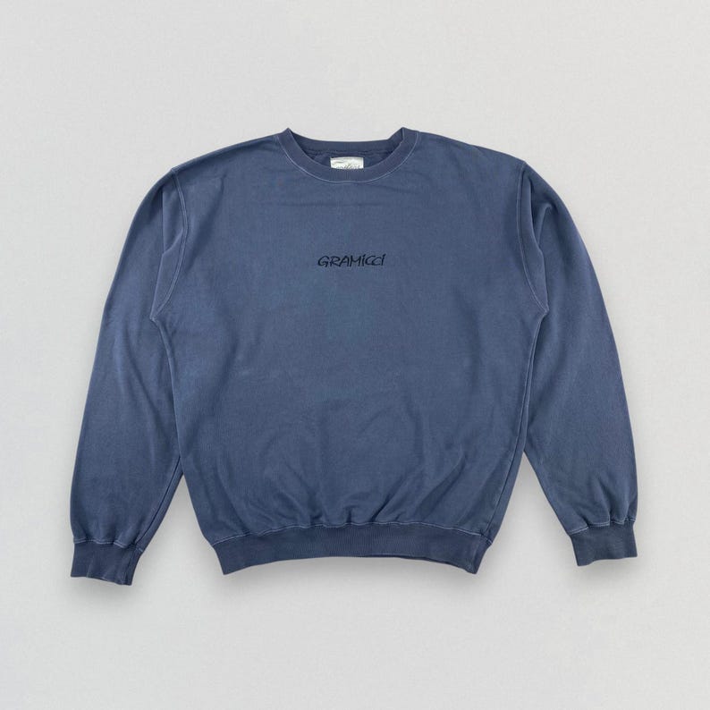 May include: A faded blue Gramicci sweatshirt with a crew neck and long sleeves. The brand name is printed in black script across the chest. The sweatshirt has a relaxed fit and a vintage aesthetic.