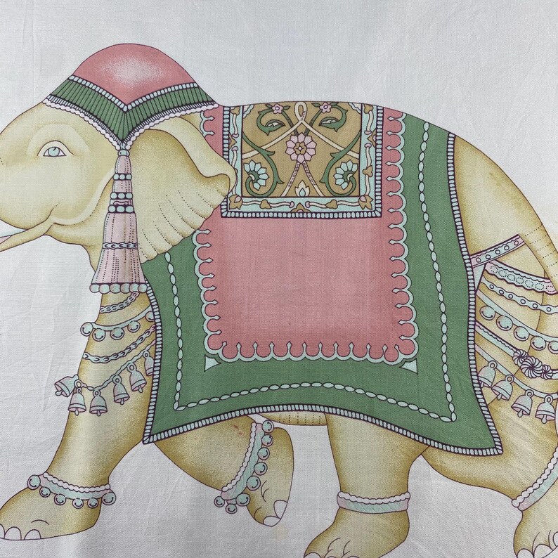May include: A printed textile featuring a stylized elephant in shades of cream, pink, and green. The elephant wears a decorative headpiece, a large saddle blanket with floral patterns, and multiple strands of hanging bells and beaded jewelry.