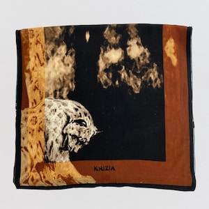 May include: Square silk scarf with a leopard design. The scarf has a black background, a brown border, and a leopard illustration. The word "KRIZIA" is printed in the bottom right corner. This scarf is a fashion accessory.