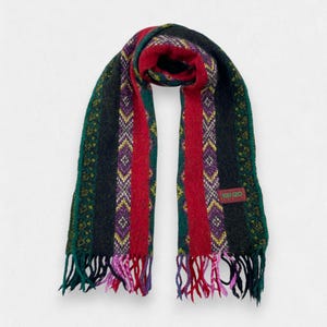 May include: A colorful scarf with a striped pattern. The scarf features vertical stripes in red, green, black, and purple, with yellow and white diamond accents. The scarf has a fringed edge and a small rectangular tag with the brand name.