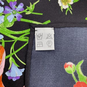 Puede incluir: Un pa&ntilde;uelo de seda negro con un estampado floral en tonos morados, azules, naranjas y verdes. El pa&ntilde;uelo tiene un borde azul oscuro y una etiqueta blanca con s&iacute;mbolos de cuidado.