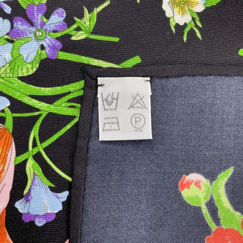 Puede incluir: Un pa&ntilde;uelo de seda negro con un estampado floral en tonos morados, azules, naranjas y verdes. El pa&ntilde;uelo tiene un borde azul oscuro y una etiqueta blanca con s&iacute;mbolos de cuidado.