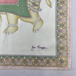 May include: A square silk scarf featuring a beige background with a central illustration of an elephant adorned in a green and pink blanket with bells. The border displays a repeating floral pattern in green, pink, and gold. The text Jim Thompson is printed.
