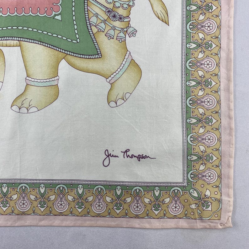 May include: A square silk scarf featuring a beige background with a central illustration of an elephant adorned in a green and pink blanket with bells. The border displays a repeating floral pattern in green, pink, and gold. The text Jim Thompson is printed.