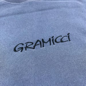 May include: A light blue crew neck sweatshirt with the embroidered word "GRAMICCI" in black. The sweatshirt has a ribbed neckline and a soft, textured appearance.