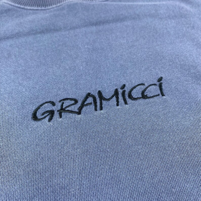 May include: A light blue crew neck sweatshirt with the embroidered word "GRAMICCI" in black. The sweatshirt has a ribbed neckline and a soft, textured appearance.