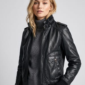 Puede incluir: Una chaqueta de cuero negra con cuello alto con correa de hebilla, puños acanalados y dos bolsillos delanteros con solapas abotonadas. La chaqueta se lleva sobre un suéter de cuello alto gris, mostrando un diseño estructurado con charreteras.