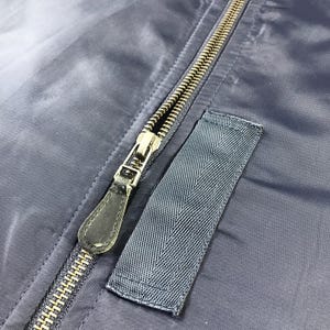 Puede incluir: Primer plano de una cremallera azul oscuro en una chaqueta. La cremallera tiene un tirador de cuero negro y est&aacute; parcialmente cerrada. La chaqueta tiene un peque&ntilde;o parche azul oscuro con una superficie texturizada.