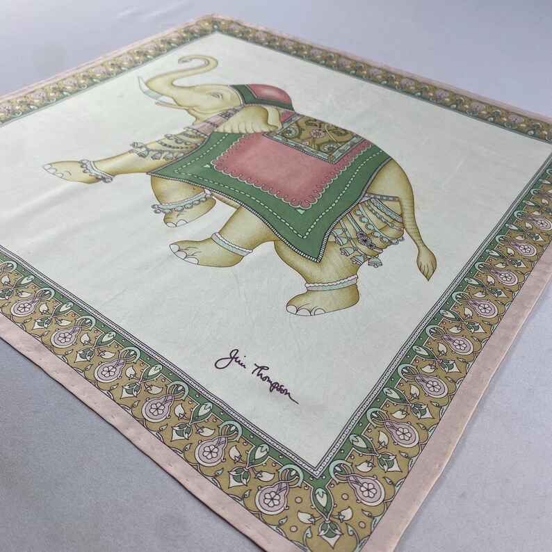 May include: A square silk scarf featuring a central illustration of an elephant adorned with a green and pink blanket and decorative leg bands. The border displays a floral pattern in shades of green, pink, and gold. The signature Jim Thompson is printed.
