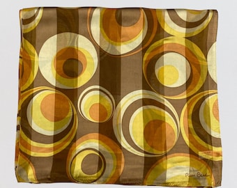 May include: A square scarf featuring a repeating pattern of concentric circles in shades of brown, orange, and yellow. The fabric has a subtle vertical striped texture throughout. The name Pierre Cardin is printed in the bottom right corner.