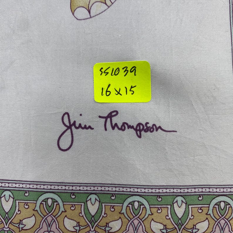 May include: A light-colored fabric featuring a decorative border with green, gold, and pink patterns. A yellow sticker displays the text 551039 and 16x15 inches. The signature Jim Thompson is printed in dark purple script below the sticker.
