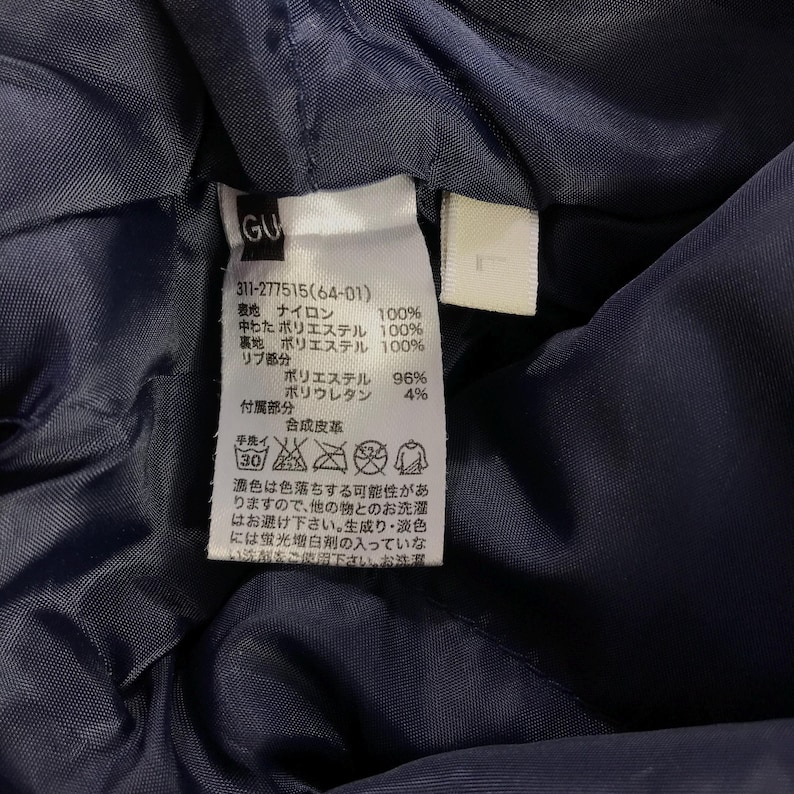 Puede incluir: Una etiqueta de ropa azul marino con texto blanco y s&iacute;mbolos. La etiqueta incluye el texto "GU", "311-277515(64-01)" y una lista de materiales e instrucciones de cuidado.