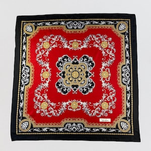 Puede incluir: Pañuelo cuadrado rojo con borde negro y detalles dorados, negros y blancos. El centro presenta un diseño simétrico con motivos florales y de volutas. El pañuelo tiene un aspecto lujoso y un estilo clásico.