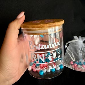 May include: A glass jar with a wooden lid, filled with pink, blue, and white beads. The jar has the name "Brianna RN 2024 Labor & Delivery" in white lettering. A small white mesh bag with beads is next to the jar.