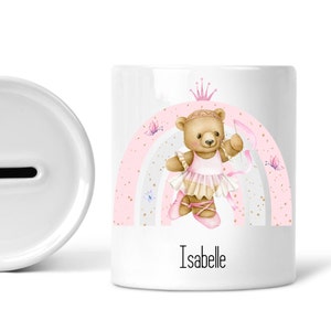 May include: White ceramic money box with a pink and gold rainbow design. A ballerina teddy bear with a crown is in the centre of the rainbow. The name "Isabelle" is written below the rainbow.