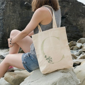 May include: A natural canvas tote bag with a large green letter "C" and green foliage. The letter is outlined in a light green colour and the foliage is a watercolour style.