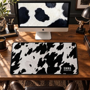 May include: A rectangular desk mat featuring a black and white cowhide print pattern. The bottom right corner displays the name EMMA in white capital letters, with the words UNTAMED SPIRIT printed in a smaller white font directly underneath.