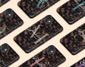 Può includere: Diverse cover rettangolari per telefono con un motivo tartarugato marrone scuro e nero. Ogni cover presenta una grande lettera maiuscola semitrasparente in un colore tenue, sovrapposta a un nome in corsivo di colore bianco.