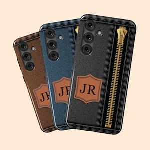 May include: Three smartphone cases with denim-like textures. The cases are brown, blue, and black, each featuring a leather patch with initials. The black case has a gold zipper detail. The cases have a camera array.