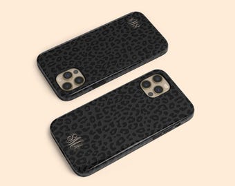 May include: Two rectangular smartphone cases featuring a black and dark gray leopard print pattern. Each case has a triple-lens camera cutout and a decorative script monogram in the corner. The cases are shown from an angled perspective.