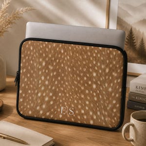 Könnte beinhalten: Eine rechteckige Laptoptasche mit einem braun-weißen Punktemuster. Die Tasche hat einen schwarzen Reißverschlussrand und ist mit den weißen Großbuchstaben E und S personalisiert, die durch eine vertikale Linie in der unteren Mitte getrennt sind.