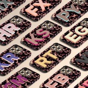 May include: A selection of smartphone cases with a leopard print pattern. Each case displays a different set of colourful 3D letters. The cases have a black border and a triple-lens camera cut-out.