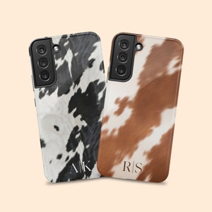 May include: Two phone cases with a cowhide pattern. One case is black and white, the other is brown and white. Both cases have initials on the back. The black and white case has the initials "AK" and the brown and white case has the initials "RS".