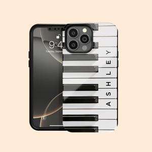 May include: A black smartphone with a white and black piano key design phone case. The case features the name "ASHLEY" in white capital letters. The phone screen displays the time as 9:41 AM on Tuesday, September.