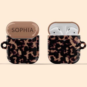 Puede incluir: Una funda para Airpods con estampado de caparazón de tortuga marrón y negro con una tapa marrón claro que dice "SOPHIA" en letras negras. La funda está abierta y los Airpods blancos son visibles en el interior.