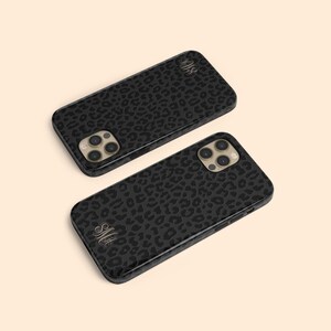 Pode incluir: Duas capas de telefone pretas com um design de estampa de leopardo e monogramas dourados. As capas são para um iPhone com uma protuberância da câmera.