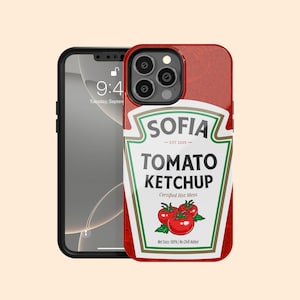 以下が含まれることがあります： トマトケチャップのラベルデザインが施された赤い携帯電話ケース。「SOFIA TOMATO KETCHUP」と書かれたラベルにはトマトのグラフィックが描かれています。携帯電話には、9時41分、9月火曜日と表示されています。