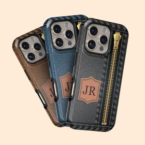 May include: Three smartphone cases with a denim effect in brown, blue, and grey. Each case has a leather patch with initials and a zipper design. The cases are designed to fit a smartphone.
