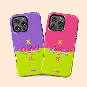 May include: Two phone cases with a vibrant design. One case is purple and green, the other is pink and green. Both cases have the word "VIBES" in a colorful font, a yellow cross, and a name at the bottom.