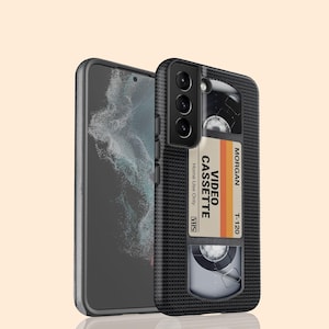 May include: A black phone case with a retro VHS cassette tape design. The cassette label reads "VIDEO CASSETTE" with the brand name "MORGAN" and "T-120". The phone case is shown next to a phone with a black screen.