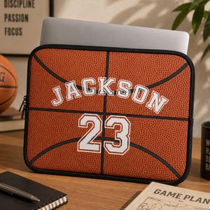 Puede incluir: Una funda rectangular para portátil con un patrón de baloncesto naranja texturizado y líneas negras. El nombre JACKSON está impreso en letras mayúsculas blancas con un borde negro, seguido del número 23 en un estilo de fuente a juego.