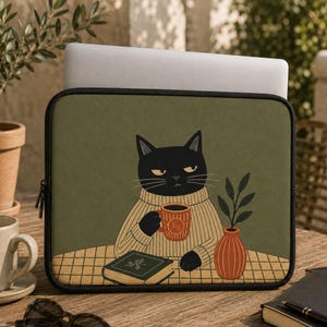 May include: A rectangular laptop sleeve with a dark olive green background featuring an illustration of a black cat wearing a beige jumper, holding a red mug with the letters ES, sitting at a table with a closed book and a small potted plant.