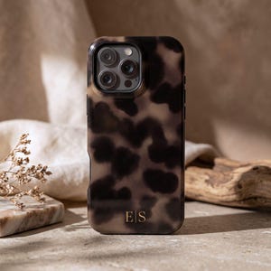 May include: A smartphone case featuring a dark brown and tan tortoiseshell pattern with a smooth finish. The case includes a cutout for a triple-lens camera system and displays the gold-colored initials E|S printed near the bottom edge.