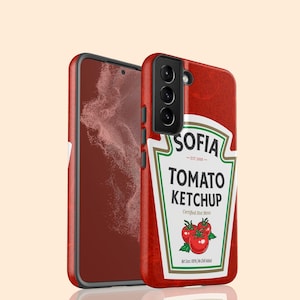 以下が含まれることがあります： トマトケチャップのデザインが施された赤いスマートフォンケース。ケースには「SOFIA TOMATO KETCHUP」の文字とトマトのグラフィックが描かれています。黒いフレームのスマートフォンと一緒に表示されています。