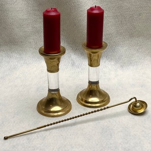 May include: Two brass candlesticks with clear acrylic stems hold red candles. A brass candle snuffer with a twisted handle lies nearby. The candlesticks have a classic design, suitable for home decor or special occasions.