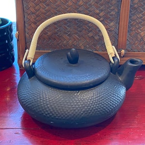 May include: A black ceramic teapot with a textured surface and a woven handle. The teapot has a lid with a small knob on top.