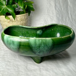 May include: A green ceramic planter with a three-footed base. The planter has a kidney bean shape and a glossy finish with varying shades of green. A potted plant with green leaves is in the background.